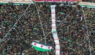 This aerial view shows people carrying a giant Syrian opposition flag before it is hoisted on a mast, during a rally to mark the 12th anniversary of the start of the uprising against Syrian President Bashar al-Assad and his government, in the rebel-held northwestern city of Idlib on March 15, 2023. (Photo by OMAR HAJ KADOUR / AFP)