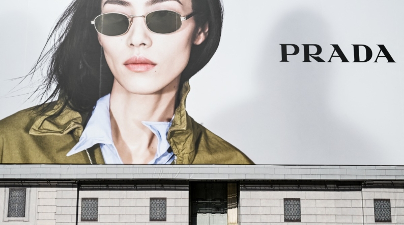 A man sits on a bench reading a newspaper next to a billboard of Italian luxury fashion brand Prada, on Navona Square on April 27, 2025. (Photo by Stefano RELLANDINI / AFP)
