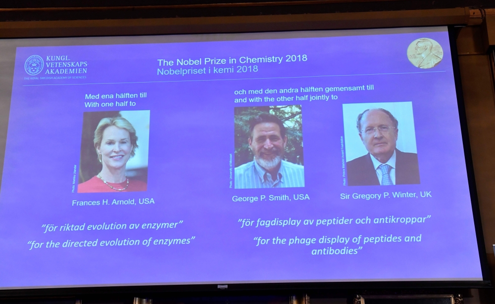 A screen displays portraits of Frances H Arnold of the United States, George P Smith of the United States and Gregory P Winter of Great Britain during the announcement of the winners of the 2018 Nobel Prize in Chemestry at the Royal Swedish Academy of Sci