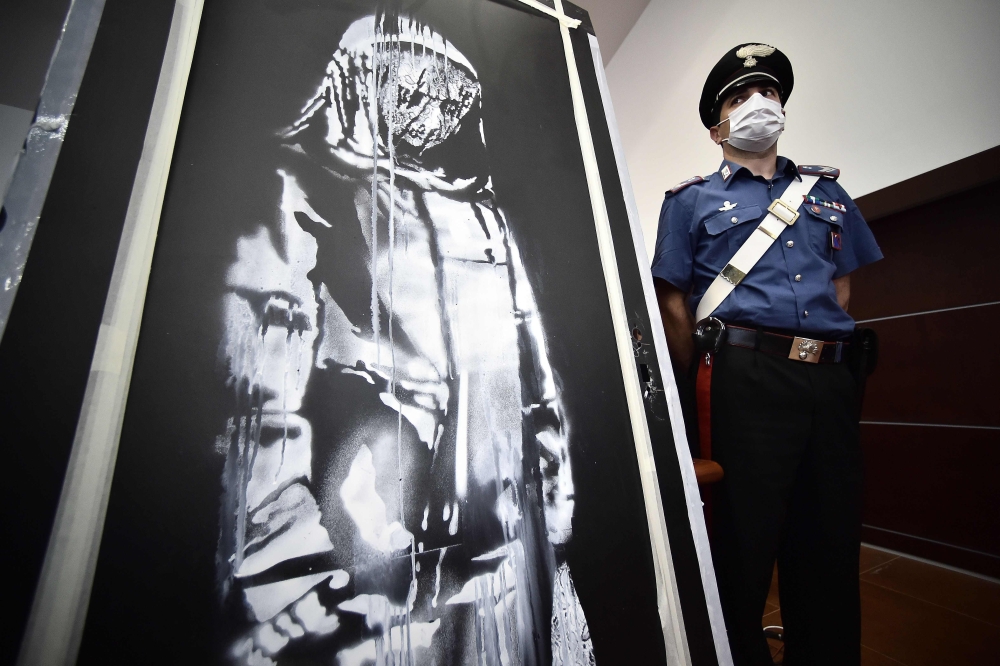 A policeman stands guard near a piece of art attributed to Banksy, that was stolen at the Bataclan in Paris in 2019, and found in Italy, ahead of a press conference in L'Aquila on June 11, 2020.  AFP / Filippo MONTEFORTE 