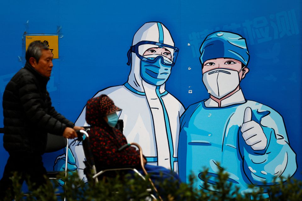 A man pushes a woman in wheelchair past a poster with a graphic of medical workers at a nucleic acid testing booth for the coronavirus disease (COVID-19) , in Beijing, China, on October 27, 2022. REUTERS/Tingshu Wang