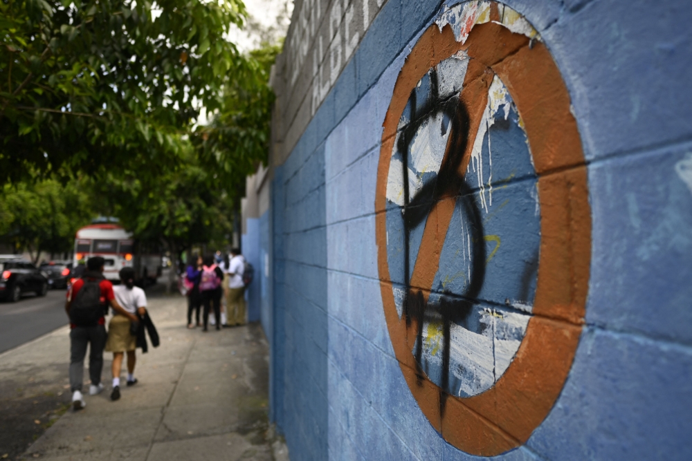 In this file photo taken on October 18, 2022, students walk past a wall painted with an anti-bitcoin protest symbol, in San Salvador. (Photo by Marvin Recinos / AFP)