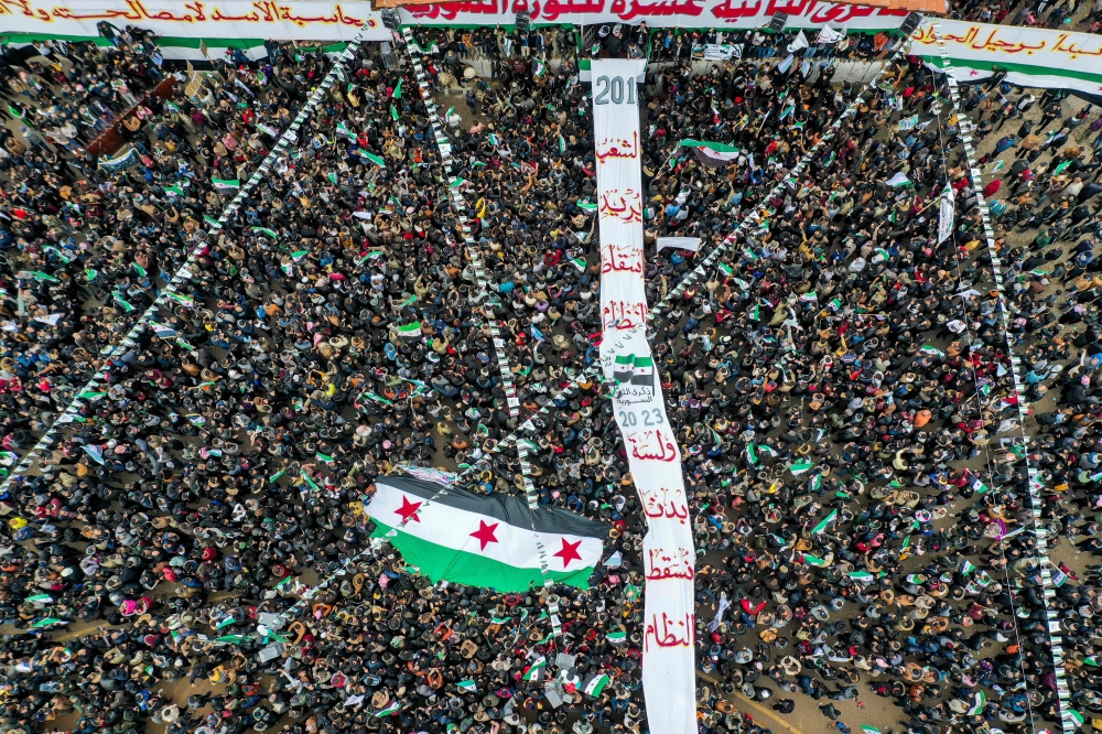 This aerial view shows people carrying a giant Syrian opposition flag before it is hoisted on a mast, during a rally to mark the 12th anniversary of the start of the uprising against Syrian President Bashar al-Assad and his government, in the rebel-held northwestern city of Idlib on March 15, 2023. (Photo by OMAR HAJ KADOUR / AFP)