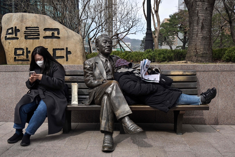A woman leans back on a statue of South Korean novelist Yeom Sang-seop to read her newspaper, as another woman uses her phone in Seoul on March 16, 2023.