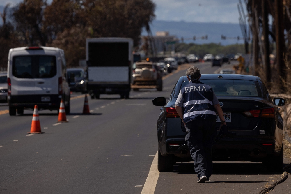 A Federal Emergency Management Agency (FEMA) agent is seen as search and recovery team members check charred buildings and cars in the aftermath of the Maui wildfires in Lahaina, Hawaii, on August 18, 2023. (Photo by Yuki IWAMURA / AFP)
