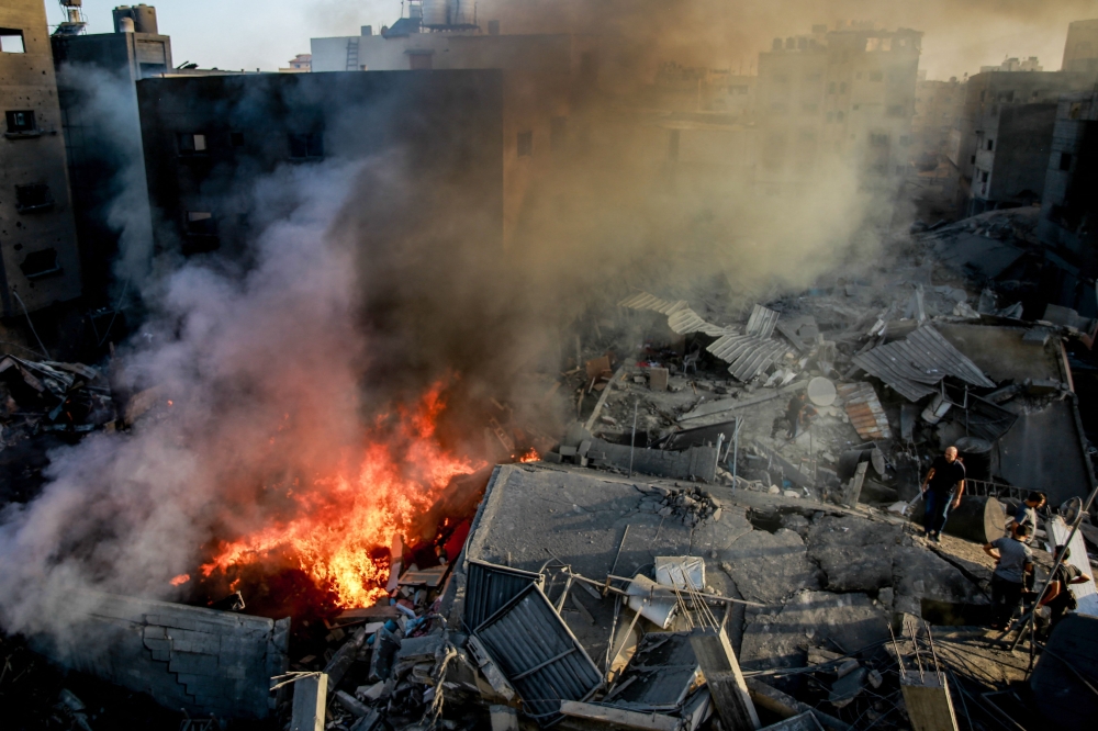 Palestinians stand on the rubble of a levelled building as smoke and fire rise from the destruction following an Israeli strike in Gaza City on October 26, 2023, amid battles between Israel and the Palestinian group Hamas. Photo by Omar El-Qattaa / AFP