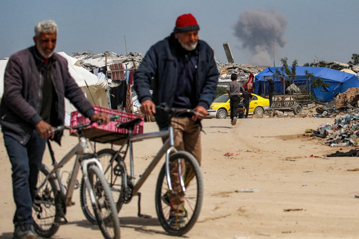 Two elderly men walk pushing bicycles while behind a cloud of smoke erupts from Israeli bombardment on the northern Gaza Strip, in Beit Lahia on March 25, 2025. Photo by Bashar TALEB / AFP