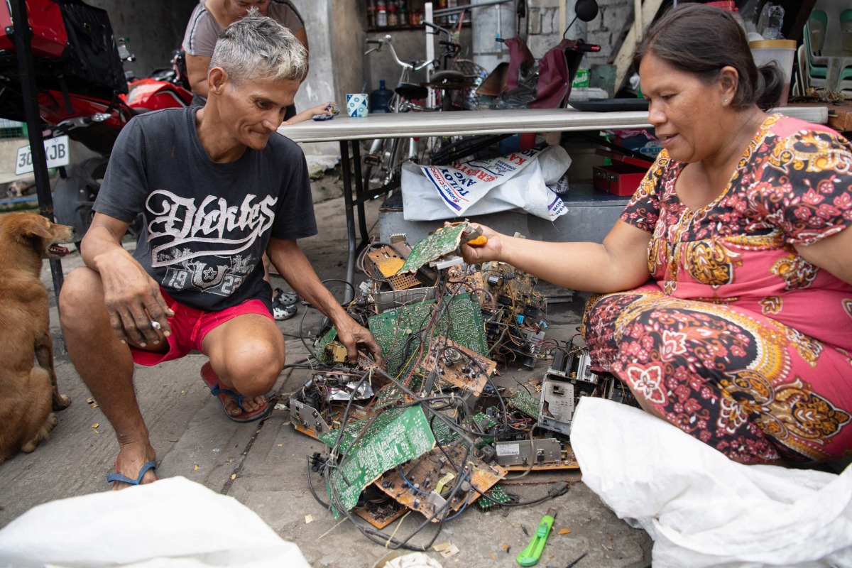 This photo taken on June 26, 2025 shows Dexter Barsigan (L), who had developed health issues after years of dismantling scavenged electronic waste, crouching beside his wife Rosalie Sedantes as she prepares to strip parts from discarded electronic equipment in Manila. (Photo by Ted ALJIBE / AFP)