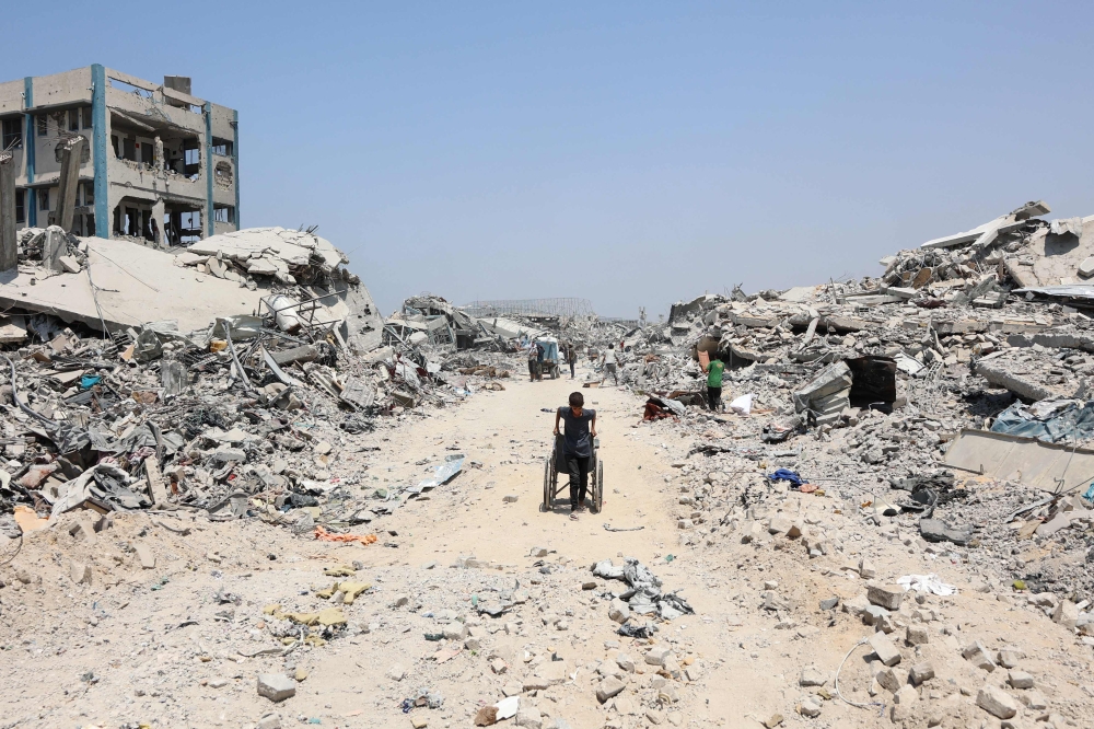 A Palestinian boy pulls a wheelchair past destroyed buildings in the al-Tuffah neighbourhood of Gaza City on August 14, 2025. (Photo by Omar Al-Qattaa / AFP)
