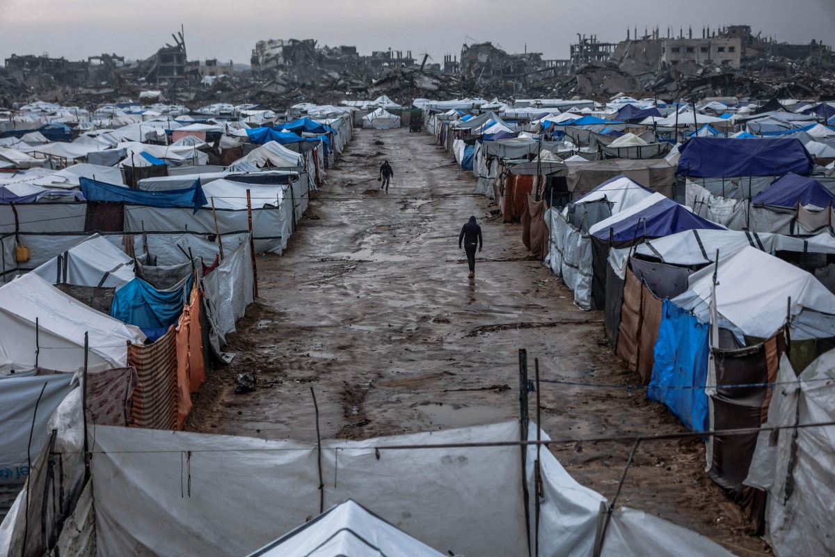 Men walk along a muddy alley at a makeshift camp sheltering displaced Palestinians after heavy rains in the Zeitoun neighbourhood of Gaza City on December 11, 2025. (Photo by Omar AL-QATTAA / AFP)