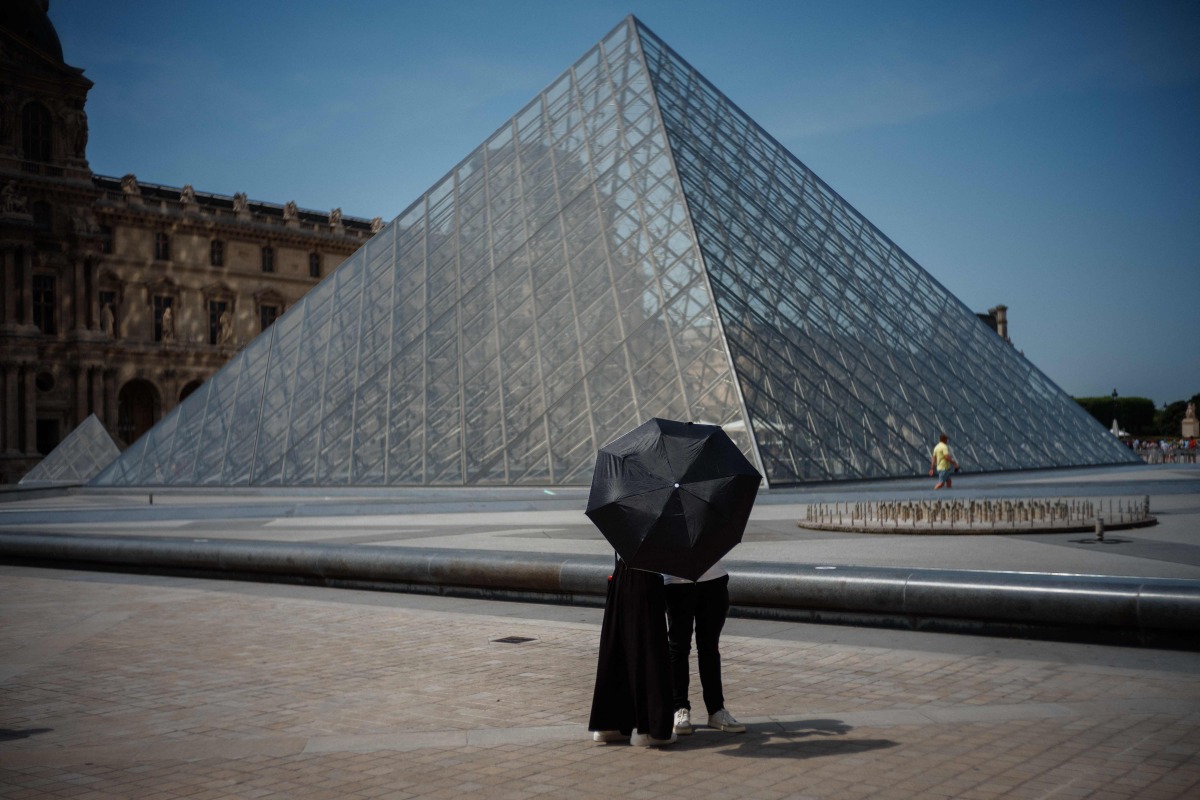 A couple protect themselves from the sun under an umbrella in front of the Louvre Pyramid, designed by Chinese-US architect Ieoh Ming Pei in Paris on June 23, 2026, as France experiences a heatwave. (Photo by Dimitar DILKOFF / AFP) 
