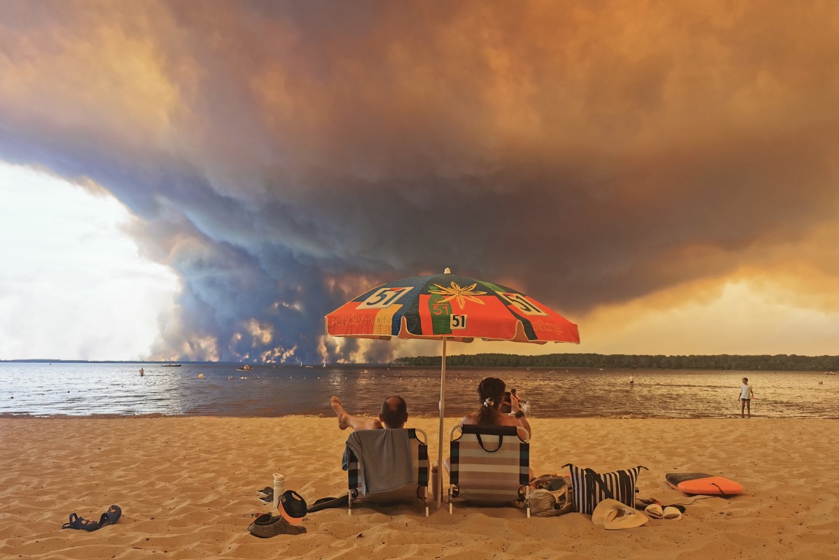 Beach goers sit on the beach of Moutchic in Lacanau, southwestern France on July 24, 2026, as clouds of smoke rise into the sky from a wildfire. Photo by Maximilien Lamy / AFP