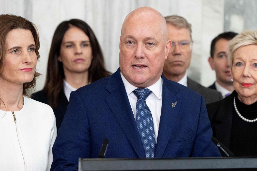 New Zealand Prime Minister Chris luxon, flanked my his caucus, speaks to the media after surviving a 2nd National party leadership vote at Parliament in Wellington on August 12, 2026. (Photo by Marty MELVILLE / AFP)