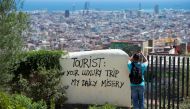 (FILES) This file photo taken on August 10, 2017 shows a tourist taking a picture of the city's panorama as he stands next to a wall with a graffito reading 'Tourist: your luxury trip - my daily misery' at Park Guell in Barcelona. While the money they bri