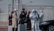 A worker (front R) wearing a protective suit as a preventive measure against the COVID-19 coronavirus stands next to passengers (L) at the New China International Exhibition Centre, near Beijing Capital Airport in Beijing on March 16, 2020./ AFP / NICOLAS