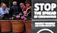 Customers sit at tables outside a restaurant, near a sign reminding members of the public to help 