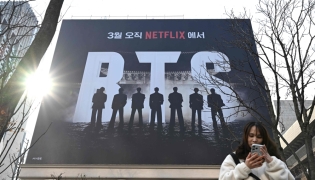 A woman uses her smartphone in front of a billboard promoting a comeback concert of K-pop boy group BTS at Gwanghwamun Square in Seoul on March 17, 2026. (Photo by Jung Yeon-je / AFP)
 