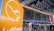 The logo of German airline Lufthansa is seen in the foreground as passengers walk below a flight information display at Frankfurt Airport, Frankfurt am Main, western Germany, on April 15, 2026, as cabin crew union UFO urged members of Lufthansa cabin crew to stage a strike in a dispute over pay and pensions. Photo by Kirill KUDRYAVTSEV / AFP