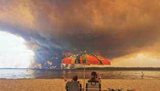 Beach goers sit on the beach of Moutchic in Lacanau, southwestern France on July 24, 2026, as clouds of smoke rise into the sky from a wildfire. Photo by Maximilien Lamy / AFP