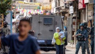 A Palestinian woman carrying a child walks past members of the Israeli occupation army and armoured vehicles during an Israeli military operation at the Balata camp for Palestinian refugees, east of Nablus in the occupied West Bank, on August 31, 2026. Photo by JAAFAR ASHTIYEH / AFP