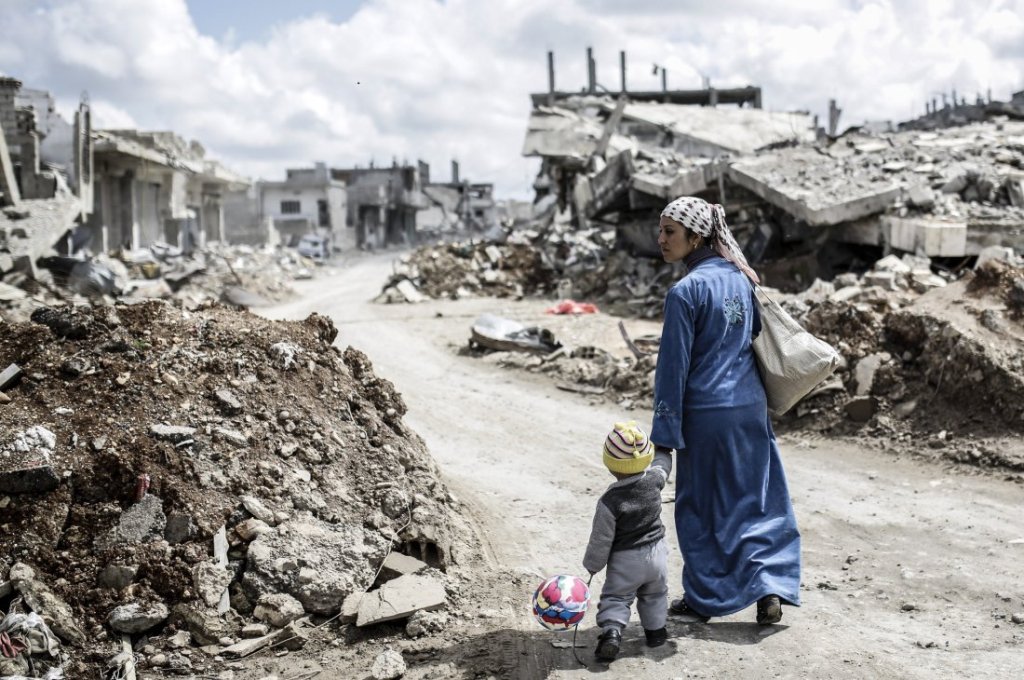 A Kurdish Syrian woman walks with her child past the ruins of the town of Kobane, also known as Ain al-Arab, on March 25, 2015. / AFP.