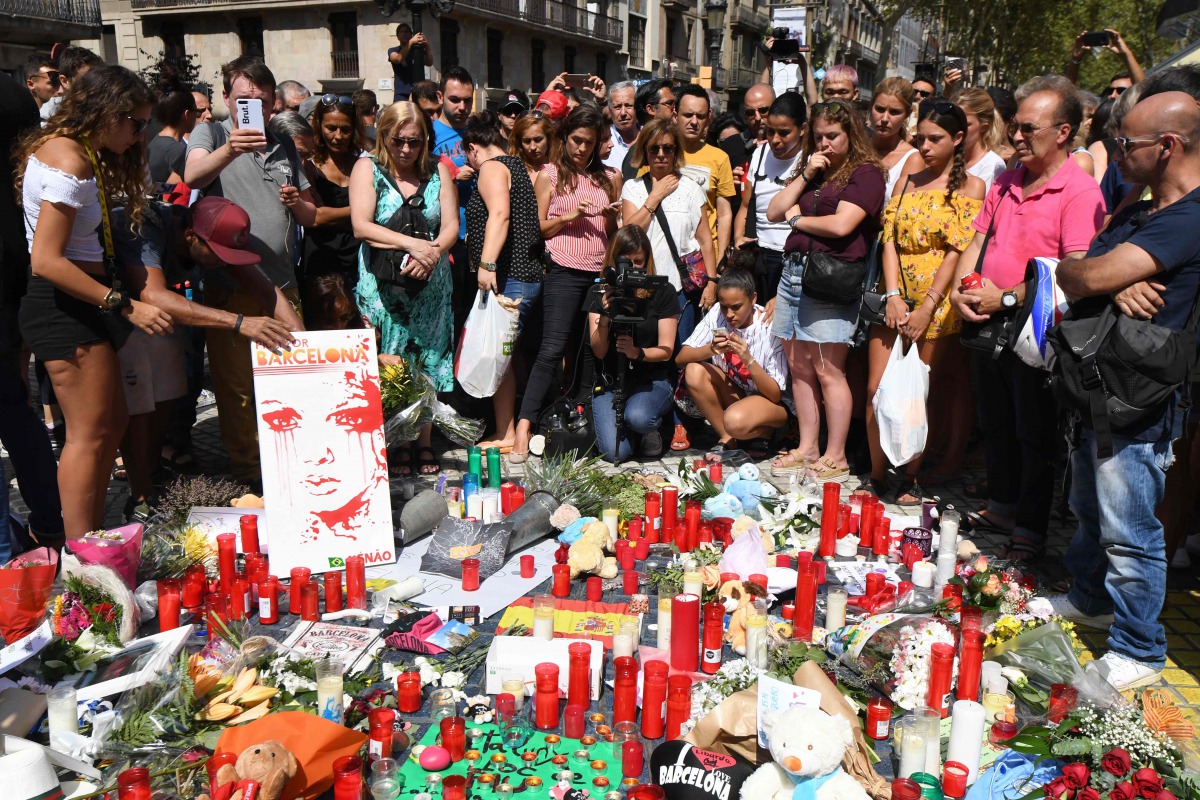People stand next to flowers, candles and other items set up on the Las Ramblas boulevard in Barcelona as they pay tribute to the victims of the Barcelona attack, a day after a van ploughed into the crowd, killing 13 persons and injuring over 100 on Augus