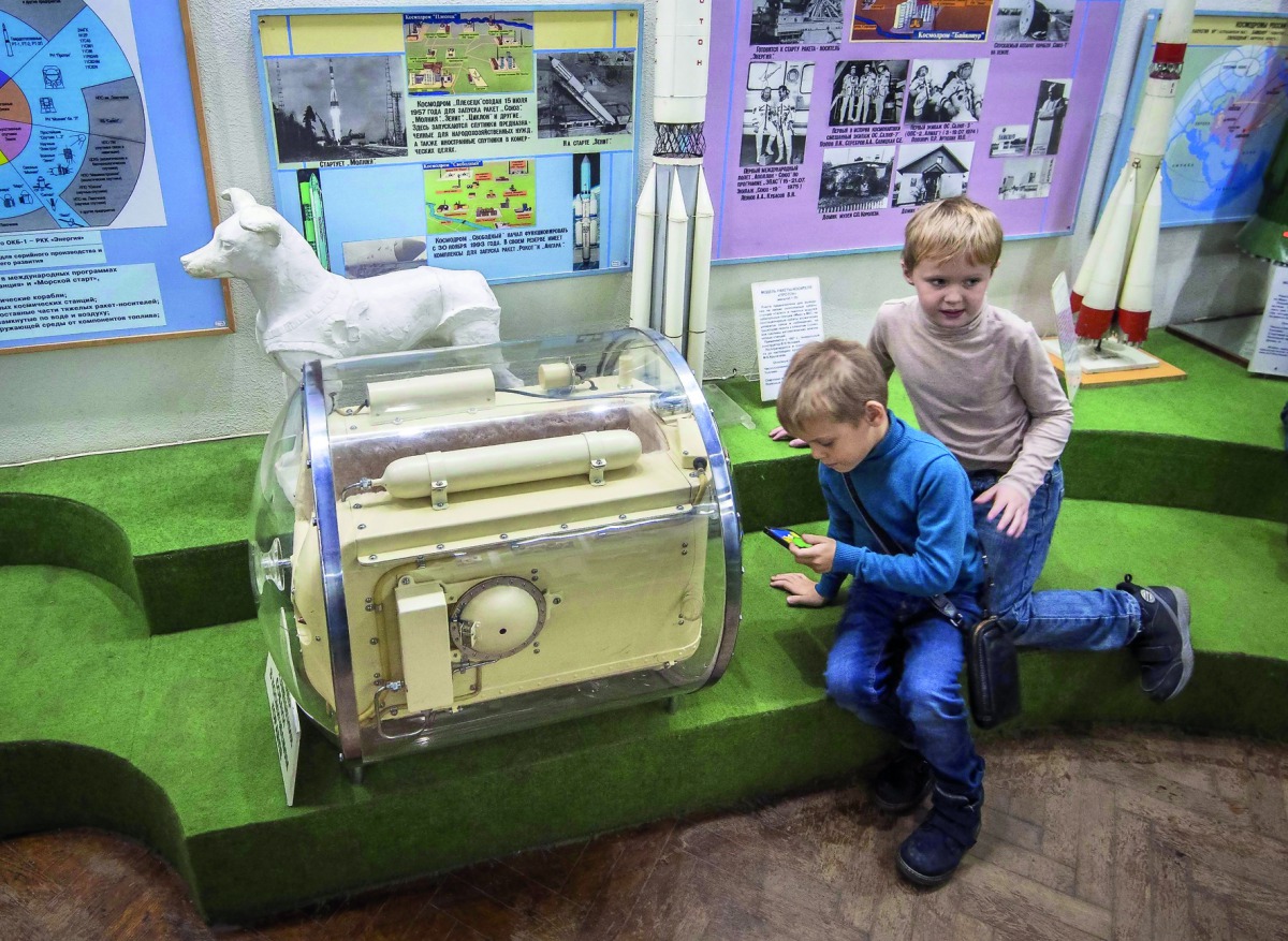 A picture taken on November 1, 2017 shows boys playing a game on a mobile phone next to the dog Laika's space container used for trainings ahead of blasting off to orbit the Earth on November 3, 1957, at the Central House of Aviation and Cosmonautics in M