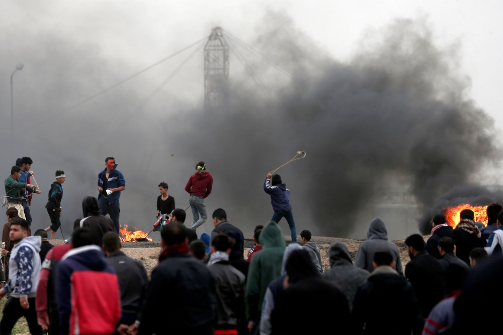 Palestinian protesters use slingshots to hurl rocks as smoke billows from tires burning during clashes with Israeli forces following a demonstration marking the first anniversary of the 