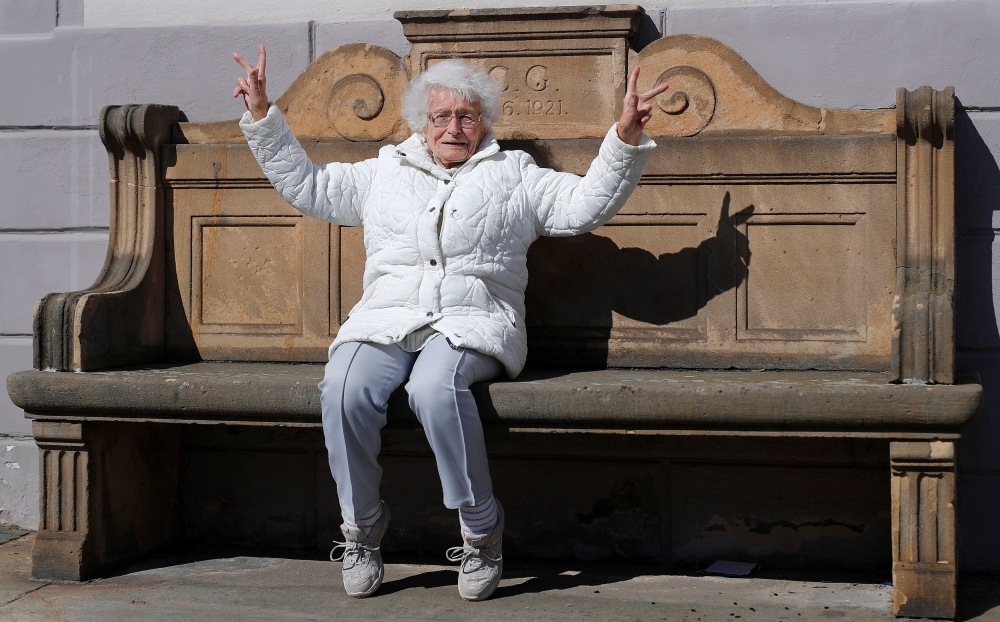Lisel Heise, a 100-year-old former teacher, sits on a stone bench from 1921 and flashes victory signs in front of the townhall in Kirchheimbolanden, Germany, April 11, 2019. REUTERS/Kai Pfaffenbach