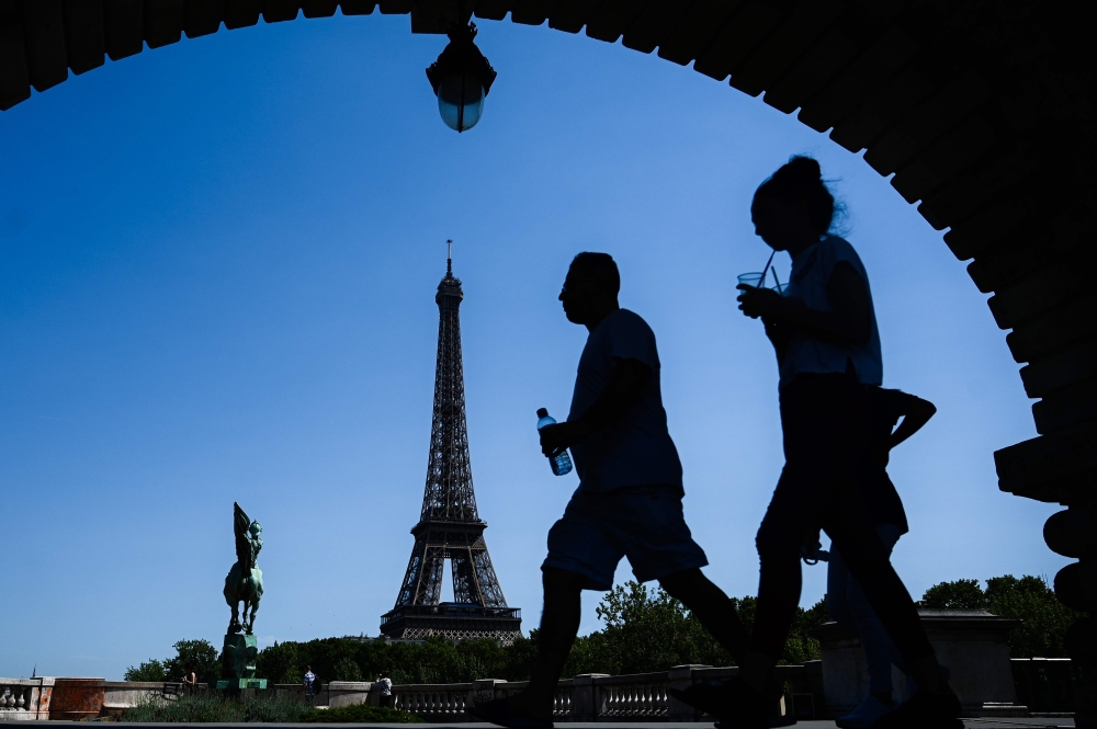 A man holding a bottle of water walks by a woman sipping a drink through a straw on a bridge over the Seine river in front of the Eiffel Tower in Paris on July 23, 2019. Parisians were bracing for potentially the hottest ever temperature in the French cap