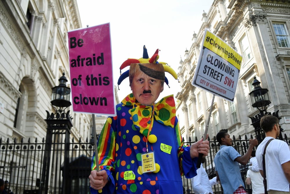 An anti-Brexit protester wearing a clown costume and a defaced mask depicting Boris Johnson holds placards in Westminster in London, Britain August 29, 2019. Reuters/Toby Melville