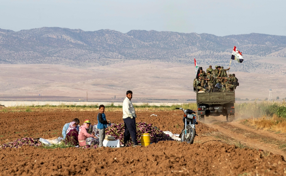 Syrian soldiers ride in the back of a truck past people sitting in a field with harvested aubergines, as government forces deploy for the first time in the eastern countryside of the city of Qamishli in the northeastern Hasakah province on November 5, 201