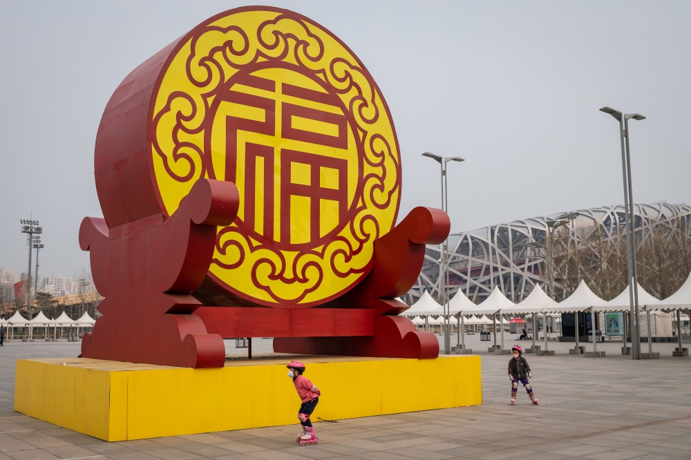 Children wearing face masks, amid concerns of the COVID-19 coronavirus, roller skate at the Olympic park outside the national 'Birds Nest' stadium (R, back), the site of the 2008 Beijing Olympics in Beijing on March 24, 2020. / AFP / NICOLAS ASFOURI