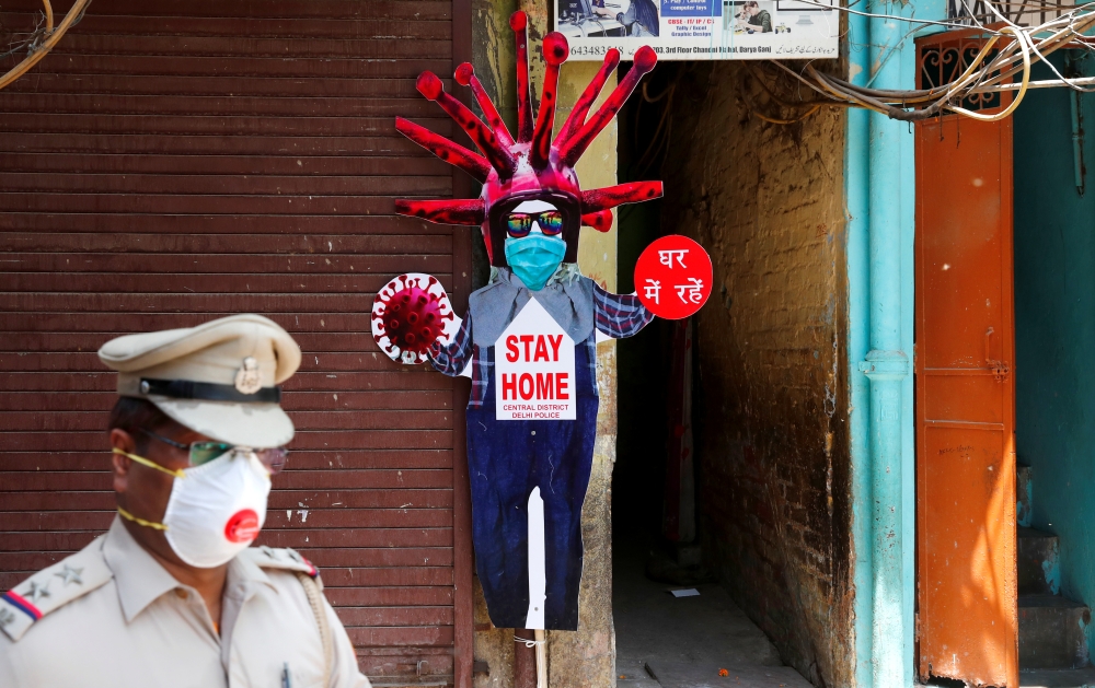 A police officer wearing a protective face mask walks past a novel coronavirus-themed scarecrow cutout put up in a residential area during an extended lockdown to slow the spreading ofthe coronavirus disease (COVID-19) in the old quarters of Delhi, India,
