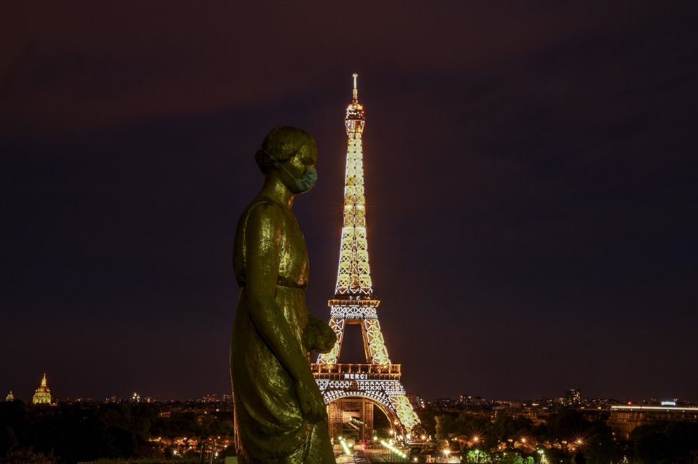 A picture taken on May 2, 2020 shows a bronze statue wearing a face mask emmulating the actions of many citizens to protecting themselves against the novel coronavirus, COVID-19 at the Parvis des Droits de l'Homme, with the Eiffel Tower enlightened in bac