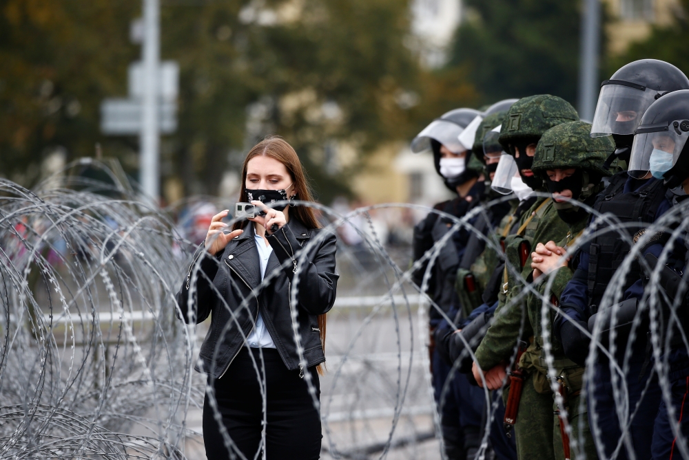A woman uses a camera next to law enforcement officers standing behind barbed wire during an opposition demonstration to protest against presidential election results, in Minsk, Belarus August 23, 2020. REUTERS/Vasily Fedosenko