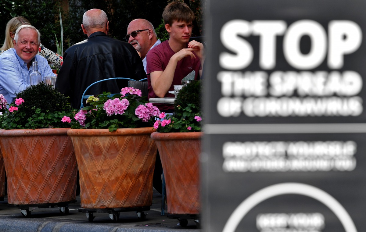 Customers sit at tables outside a restaurant, near a sign reminding members of the public to help 