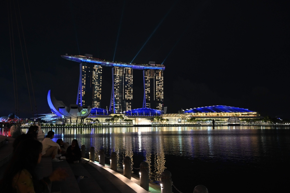 People sit along the steps of the quay watching lights illuminate Marina Bay Sands hotels and resorts to celebrate its tenth anniversary and usher in the holiday season in Singapore on November 26, 2020. / AFP / Roslan Rahman