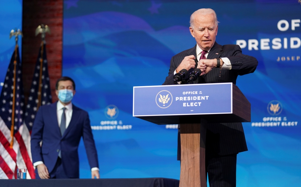 US President-elect Joe Biden looks at his watch as former South Bend, Indiana Mayor Pete Buttigieg arrives behind him to be annouced as his nominee for secretary of transportation during a news conference at Biden's transition headquarters in Wilmington, 