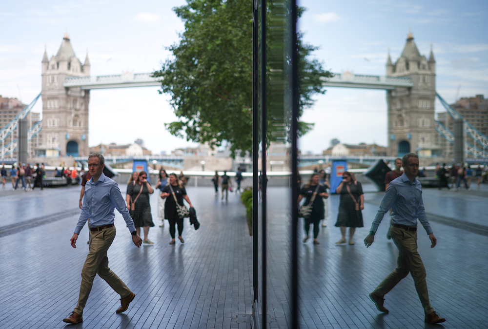 A man, reflected in a building, walks on the Southbank of the River Thames, with London Bridge on the background , central London, on August 14, 2023. Photo by HENRY NICHOLLS / AFP