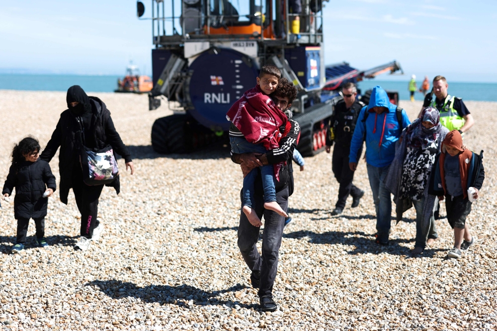 A man carrying a child in his arms, is followed by a migrant pregnant woman (2nd R), as they walk on the beach at Dungeness on the southeast coast of England, on August 16, 2023. (Photo by HENRY NICHOLLS / AFP)
 