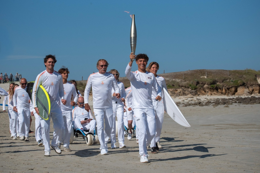 French surfer Louka Tirilly de Vera (centre) holds the Olympic flame flanked by members of the French Olympic surfing team as part of the Olympic Relay run in Plomeur, western France on June 7, 2024, ahead of the Paris 2024 Olympic and Paralympic Games. (Photo by Oscar Chuberre / AFP)

