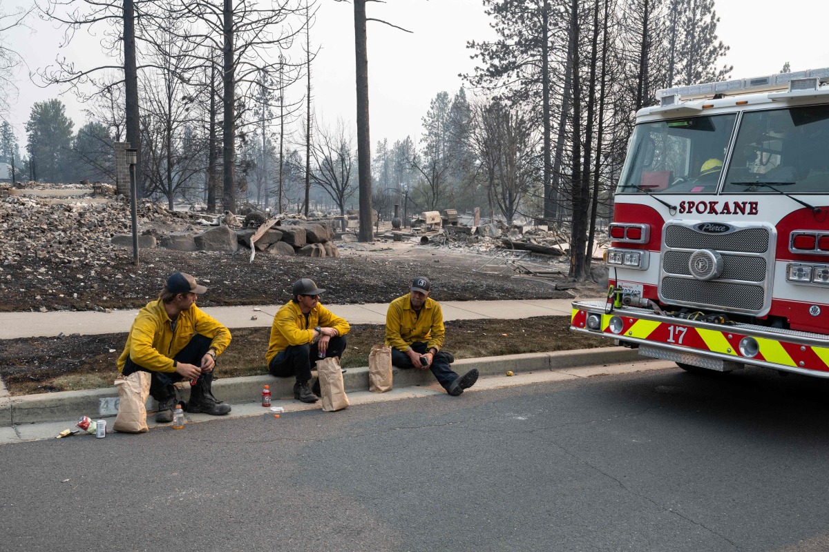 Three members of the Spokane Fire Department sit on a curb in front of burned out residential homes in the Balboa neighborhood of Spokane, Washington, on August 2, 2026. (Photo by Erick Doxey / AFP)